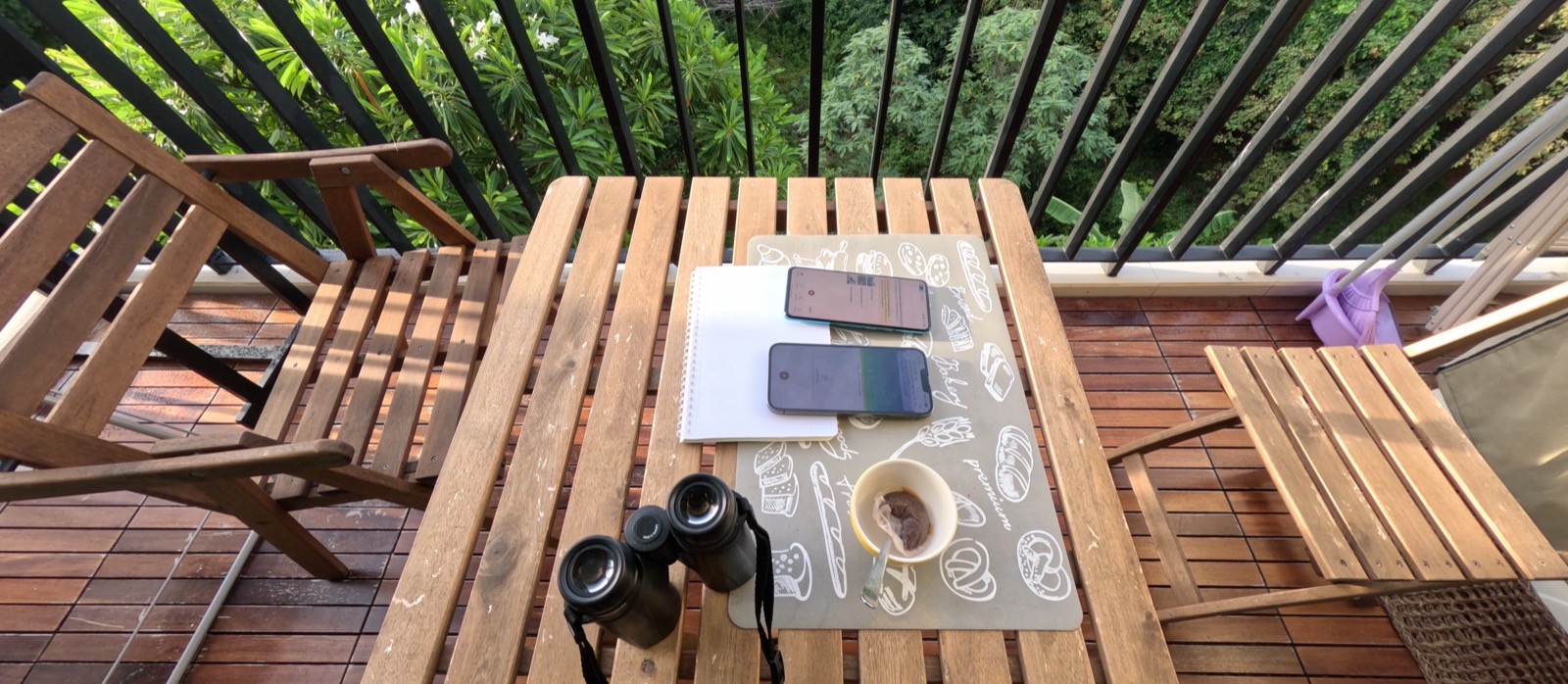 Vue plongeante sur la table du balcon : deux téléphones côte à côte sur un set de table, un carnet, une paire de jumelles et un café